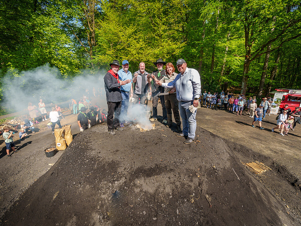 Vier PErsonen stehen auf dem Holzkohlemeilers mit einem Glas in der Hand und blicken in Richtung Kamera.