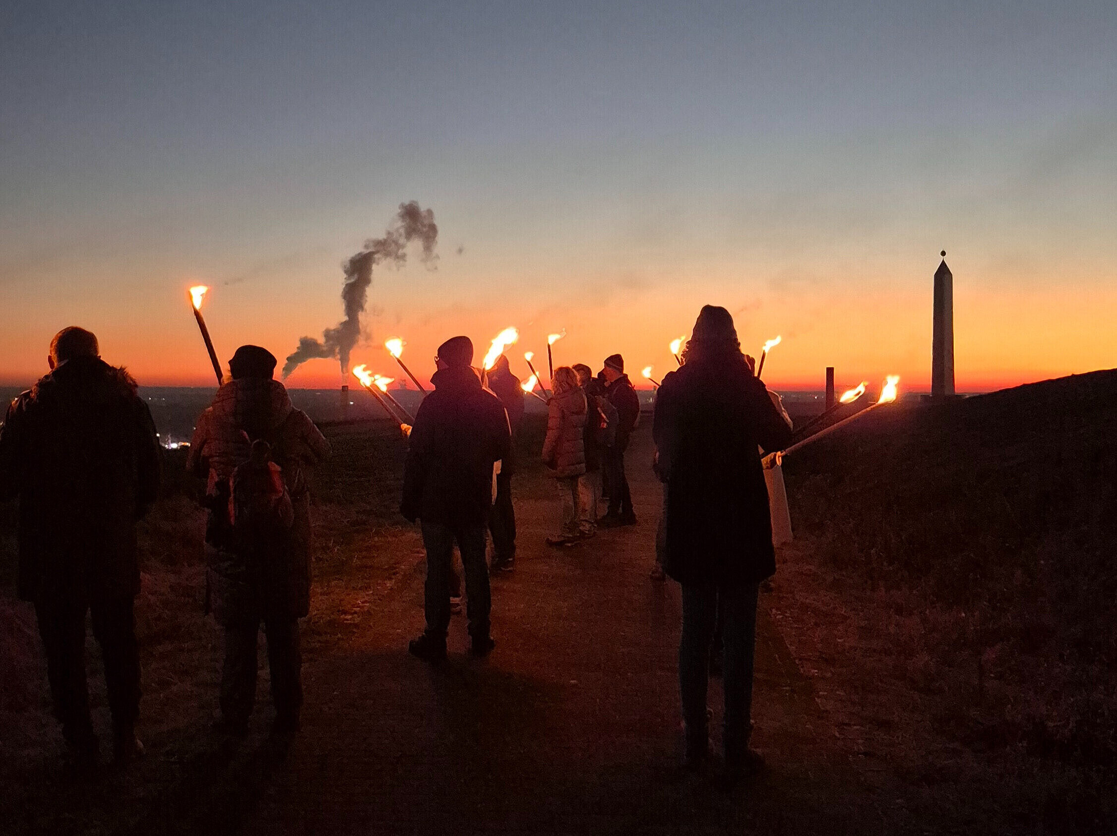 Eine Gruppe Menschen mit Fackeln läuft im Sonnenuntergang Richtung Sonnenuhr der Halde Hoheward.