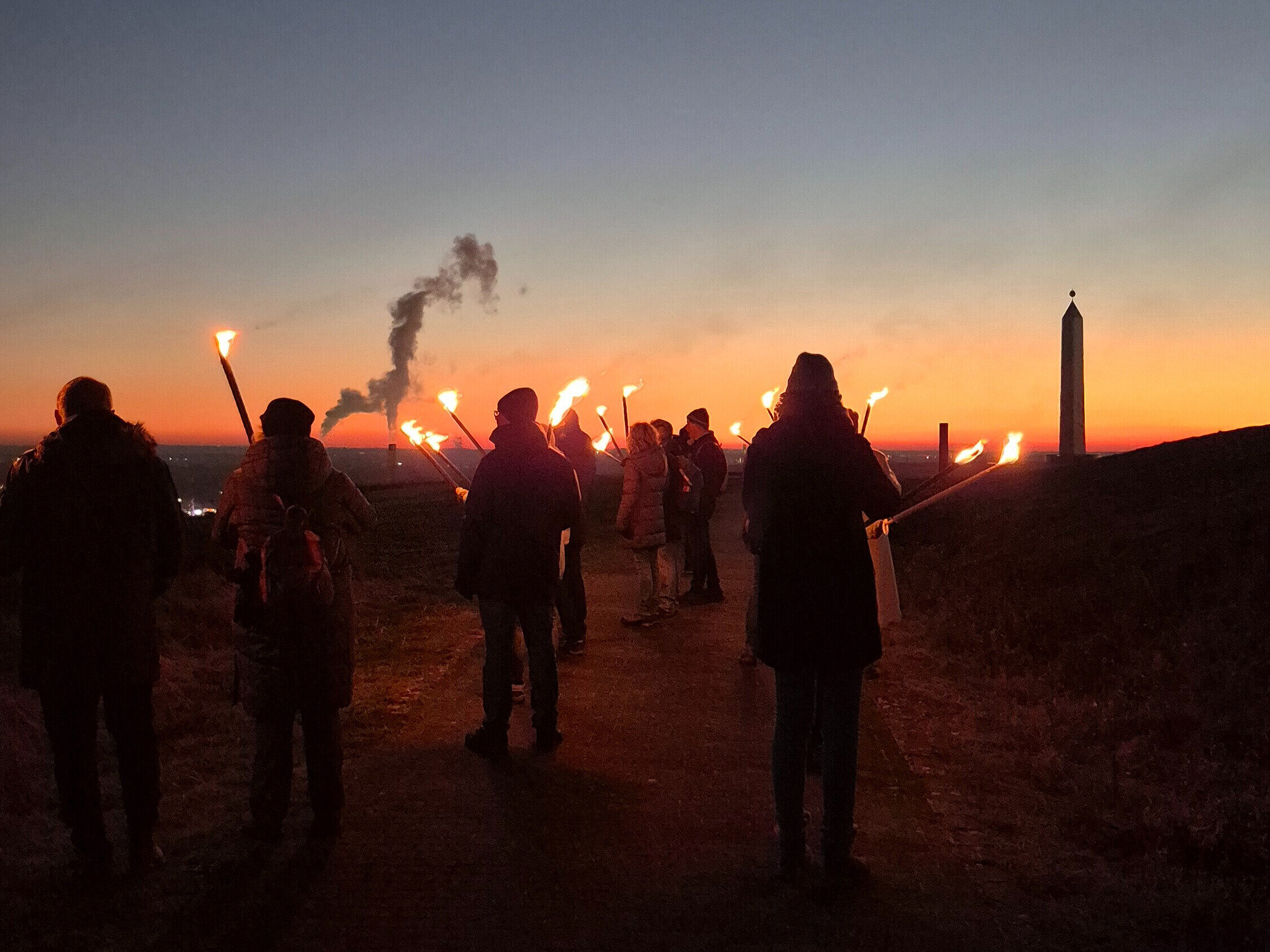 Eine Gruppe Menschen mit Fackeln läuft im Sonnenuntergang Richtung Sonnenuhr der Halde Hoheward.