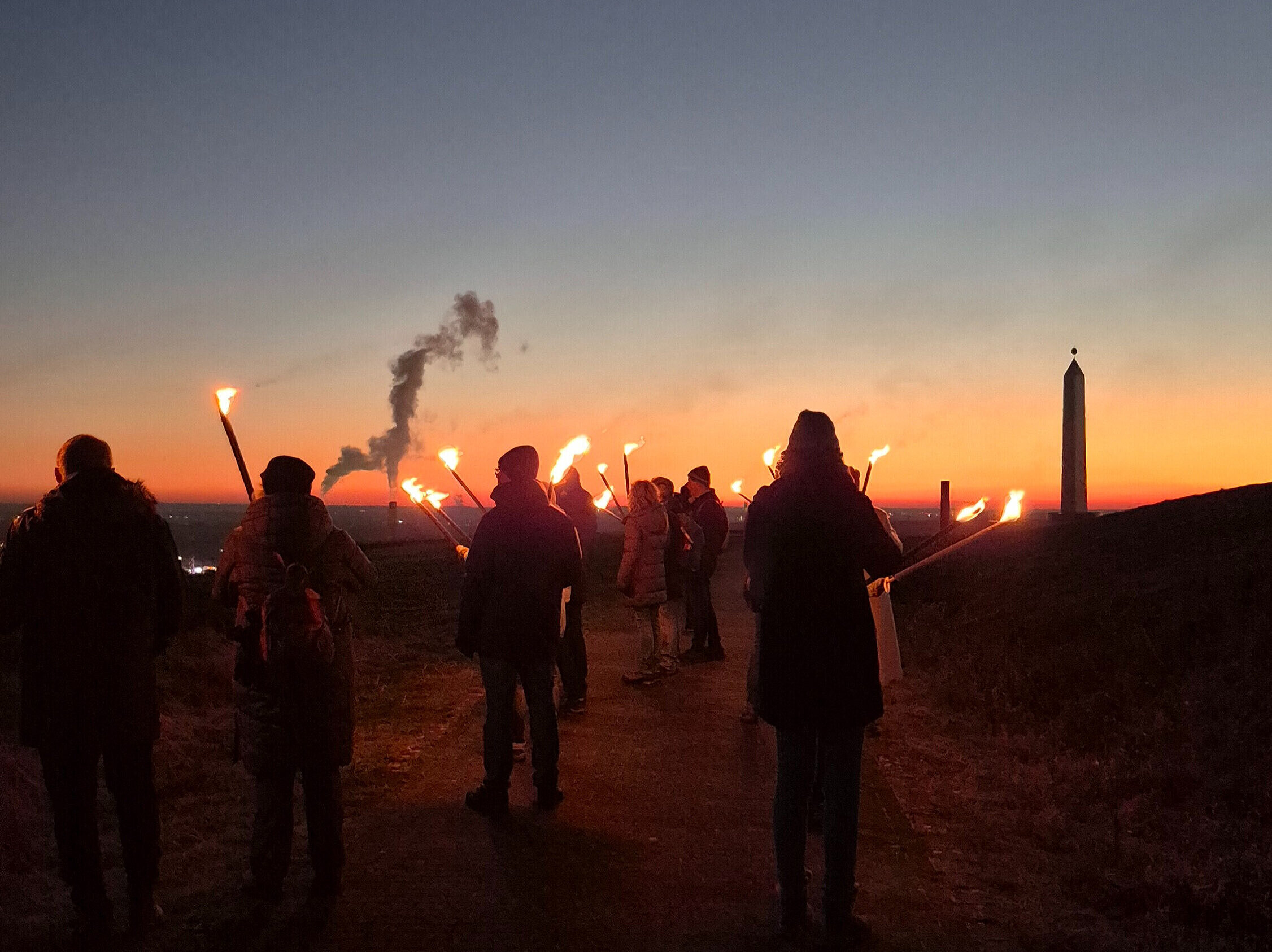 Eine Gruppe Menschen mit Fackeln läuft im Sonnenuntergang Richtung Sonnenuhr der Halde Hoheward.