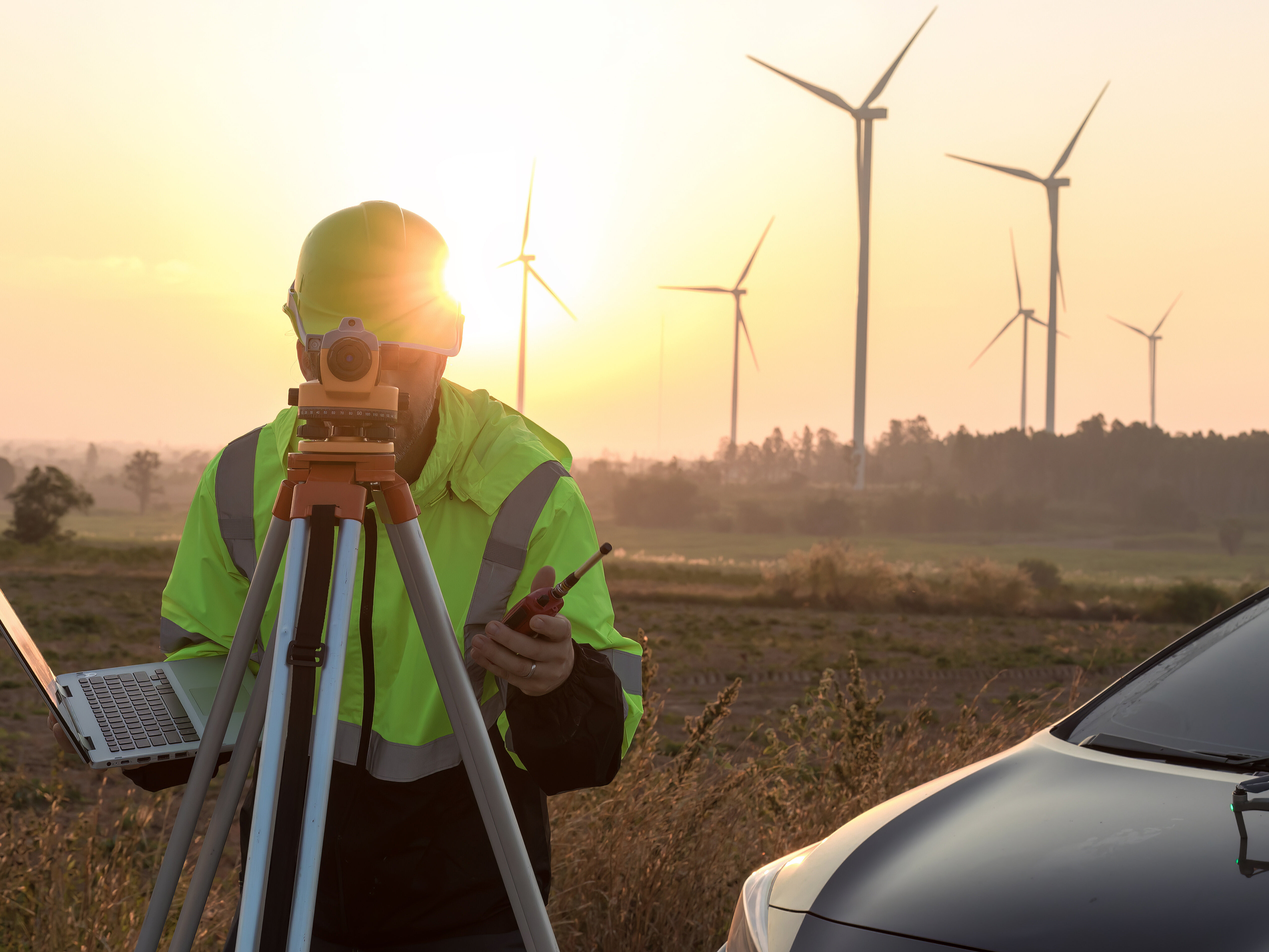 Ein Vermessungsingenieur (mit Handy, Drohne und Laptop ausgestattet) untersucht die im Hintergrund befindlichen Windturbinen im Sonnenuntergang. Rechts im Bild befindet sich ein Auto.