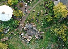 Luftbildaufnahme vom Gemeinschaftsgarten. Mittig ist eine Feuerstelle mit Bänken, an der Menschen stehen und sitzen.