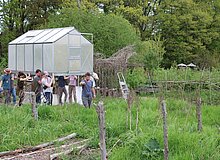 Acht Menschen schultern ein Gewächshaus und tragen es durch den Garten.