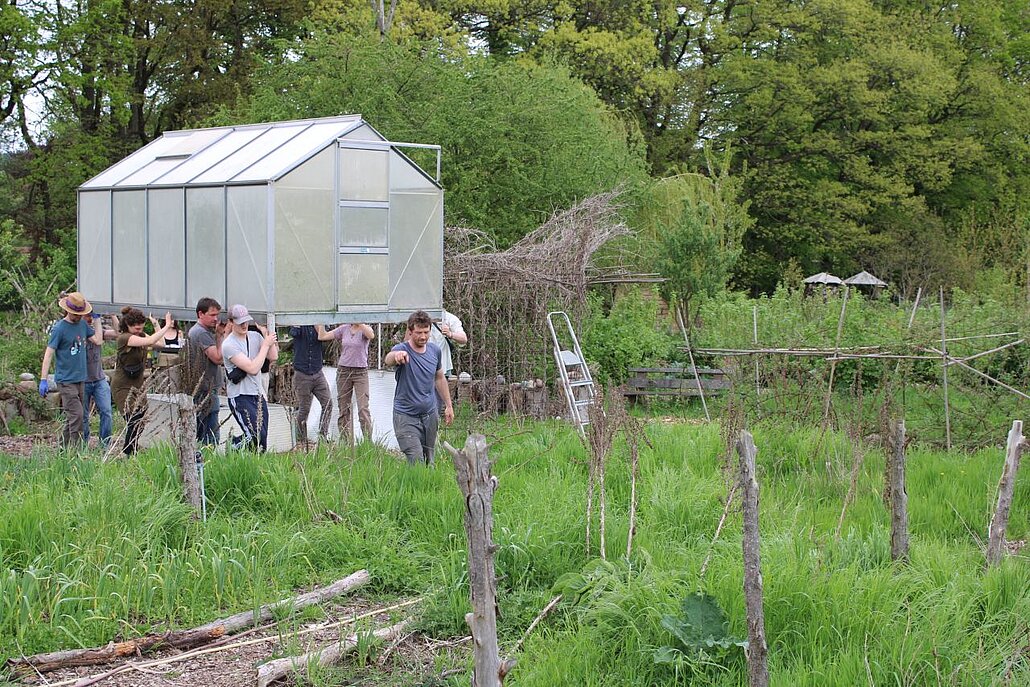 Acht Menschen schultern ein Gewächshaus und tragen es durch den Garten.