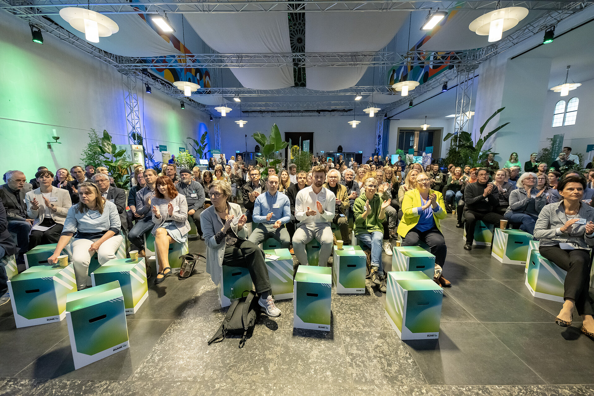 Blick von der Bühne aus ins Plenum, Teilnehmende, die auf den Sitzhockern sitzen