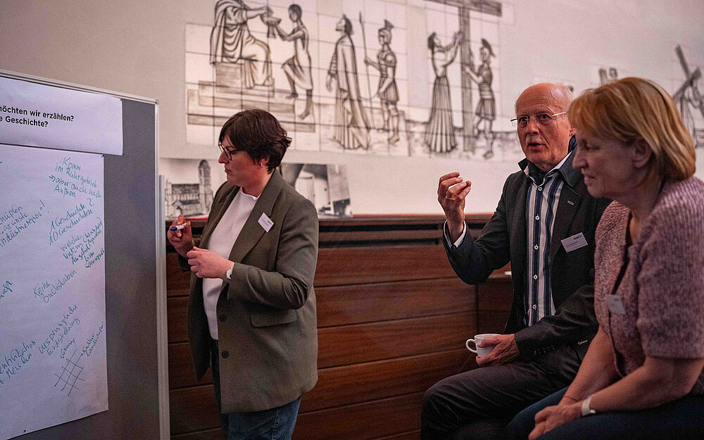 3 Personen stehen vor einem Poster. Eine Person schuat konzentert auf das Poster. Die anderen beiden unterhalten sich.