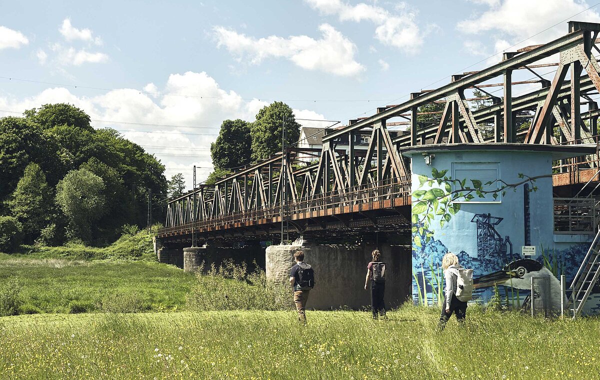 Drei Personen wandern auf einer grünen Wiese entlang einer Bahnstrecke.