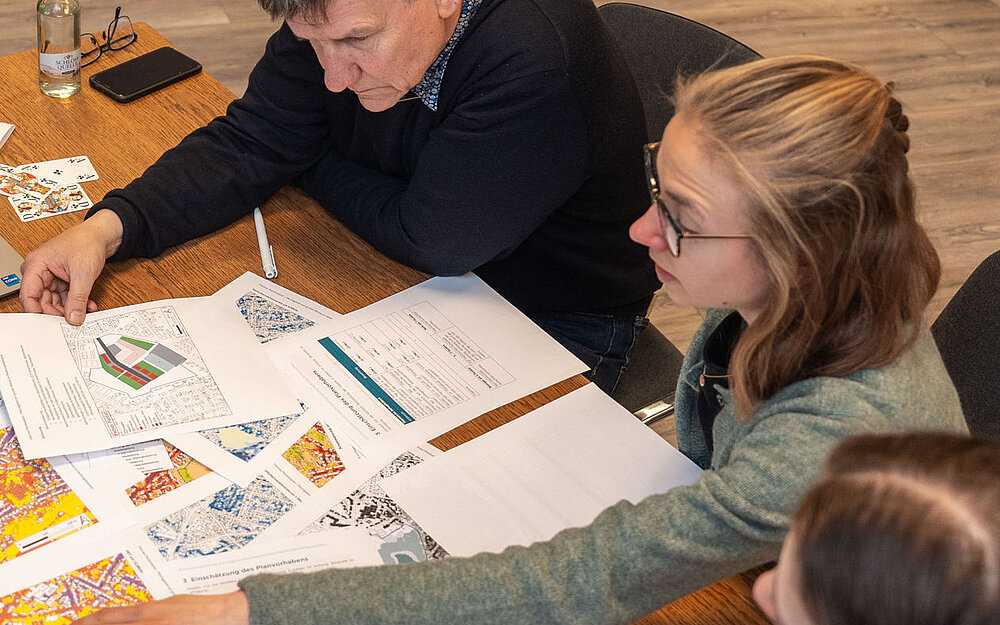 Vier Personen sitzen an einem Tisch und schauen sich auf Papier gedruckte Karten an, die auf dem Tisch liegen.