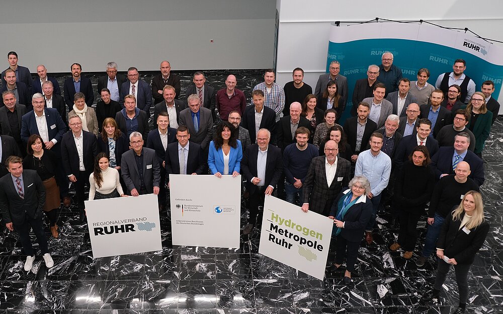 Klimaschutznetzwerk Wasserstoff Gruppe von ca. 50 Personen. Personen halten drei Schilder mit Logo des Ministeriums, RVR und Hydrogen Metropole Ruhr hoch