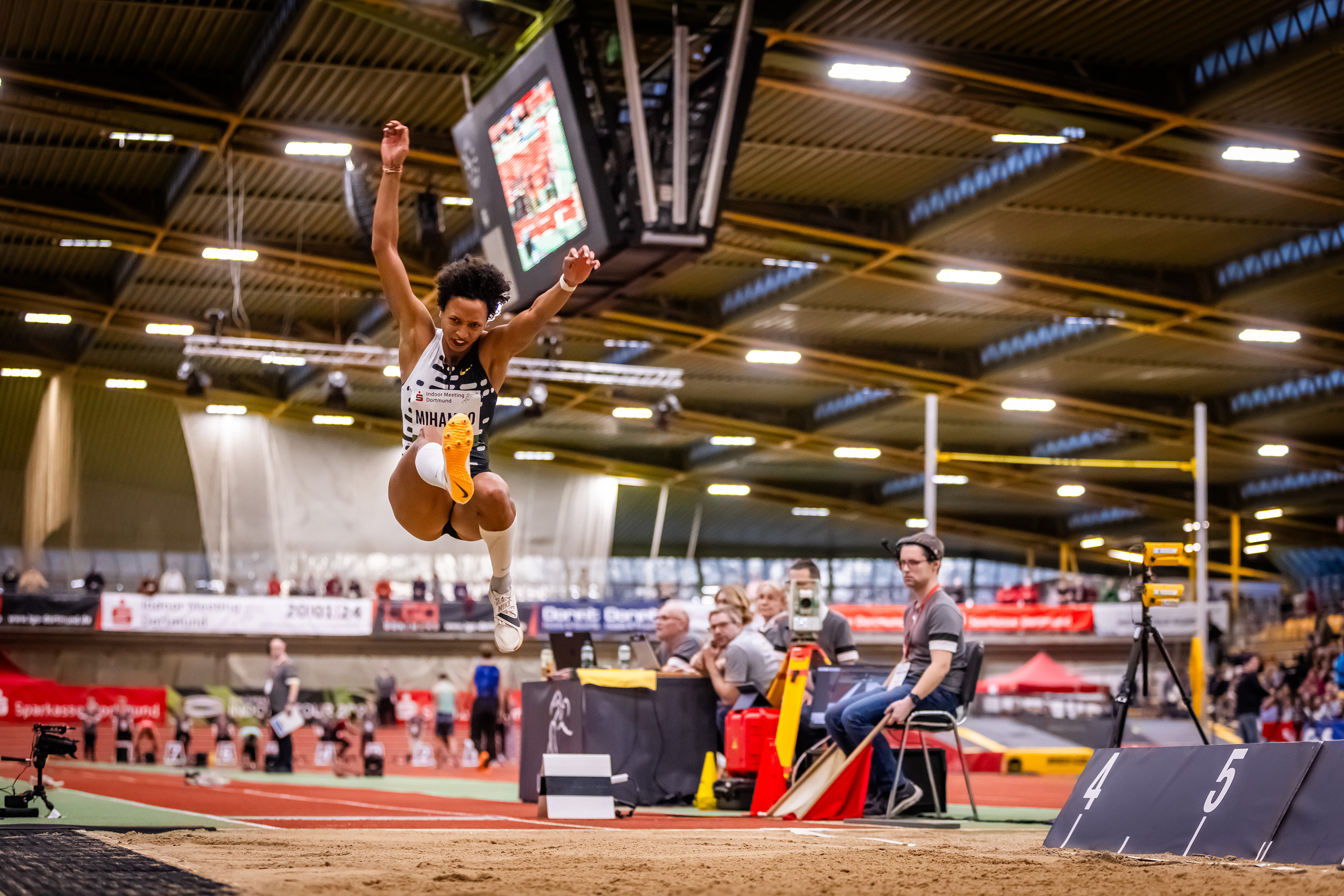 Sportlerin springt über eine Hürde in einer Sporthalle. 