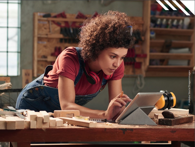 Das BIld zeigt eine Frau, die sich auf einen Tisch lehnt und am Tablet arbeitet. Sie trägt einen Arbeitsanzug aus dem Handwerk. 