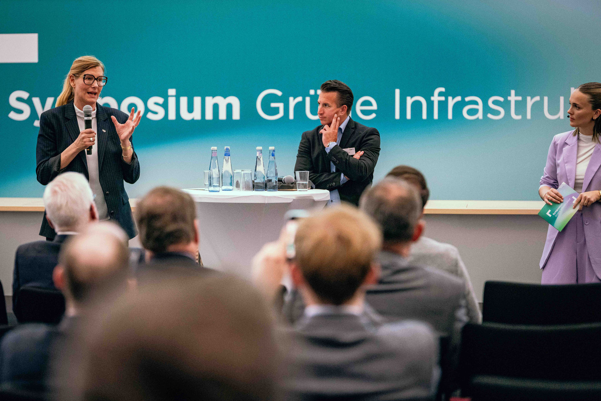 Nina Frense, Beigeordnete für den Bereich Umwelt und Grüne Infrastruktur im Regionalverband Ruhr, Christian Mildenberger, Geschäftsführer von NRW.Energy4Climate, und Moderatorin Sina Kuipers auf der Bühne des Symposiums Grüne Infrastruktur, im Vordergrund Publikum