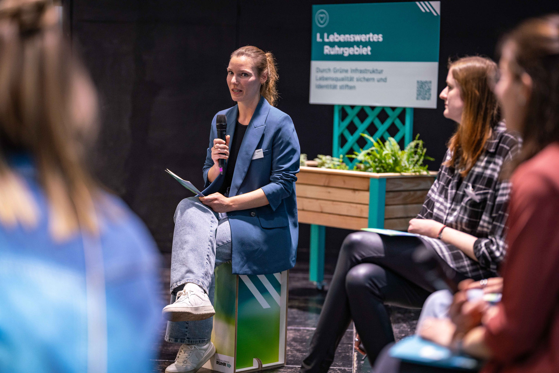 Laura Bellenberg, zuständig für Kommunikation und Veranstaltungen im Team Grüne Infrastruktur und Klimaneutralität im Referat Klima und Umweltschutz des Regionalverbands Ruhr, sitzt auf der Bühne des Netzwerktags Grüne Infrastruktur und hält ein Mikrofon in der Hand.