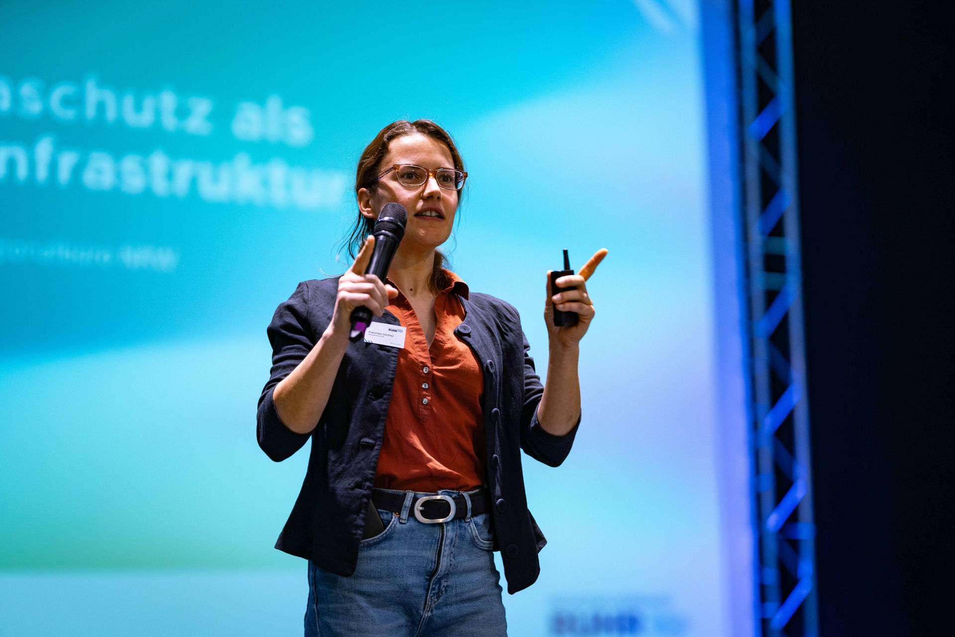 Franziska Günther vom Regionalbüro Nordrhein-Westfalen des Aktionsprogramms Natürlicher Klimaschutz steht auf der Bühne des Netzwerktags Grüne Infrastruktur und hält ein Mikrofon in der Hand.