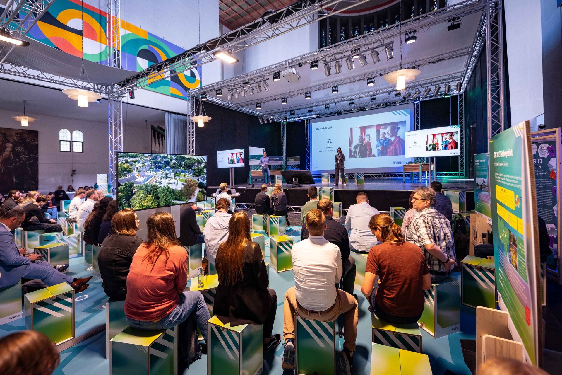  Publikum des Netzwerktags Grüne Infrastruktur auf Papphockern sitzend von hinten, im Hintergrund eine Bühne.