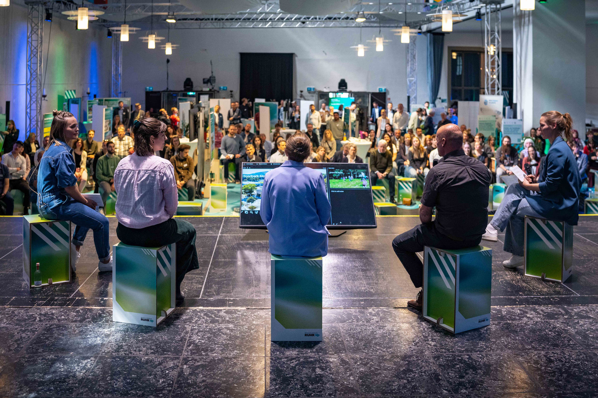 Fünf Personen sitzen auf Papphockern auf einer Bühne, sie sind von hinten fotografiert, im Hintergrund der Saal mit dem Publikum.