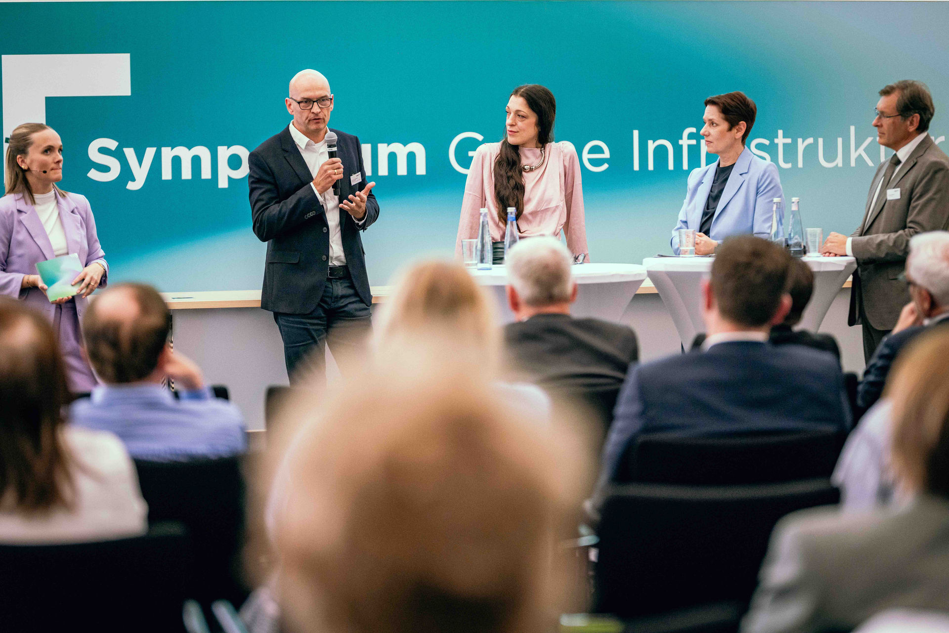 Moderatorin Sina Kuipers, Dr. Kai Gerhard Steinbrich, Geschäftsführer der ENNI Energie & Umwelt Niederrhein GmbH, Paula Backhaus, Vorsitzende des Regionalverbands Düsseldorf/Ruhr des Landesverbands Erneuerbare Energien Nordrhein-Westfalen, Silke Gorißen, Ministerin für Landwirtschaft und Verbraucherschutz des Landes Nordrhein-Westfalen, und Dr. Frank Dudda, Oberbürgermeister der Stadt Herne und Vorsitzender des Ruhrparlaments, auf der Bühne des Symposiums Grüne Infrastruktur, im Vordergrund Publikum