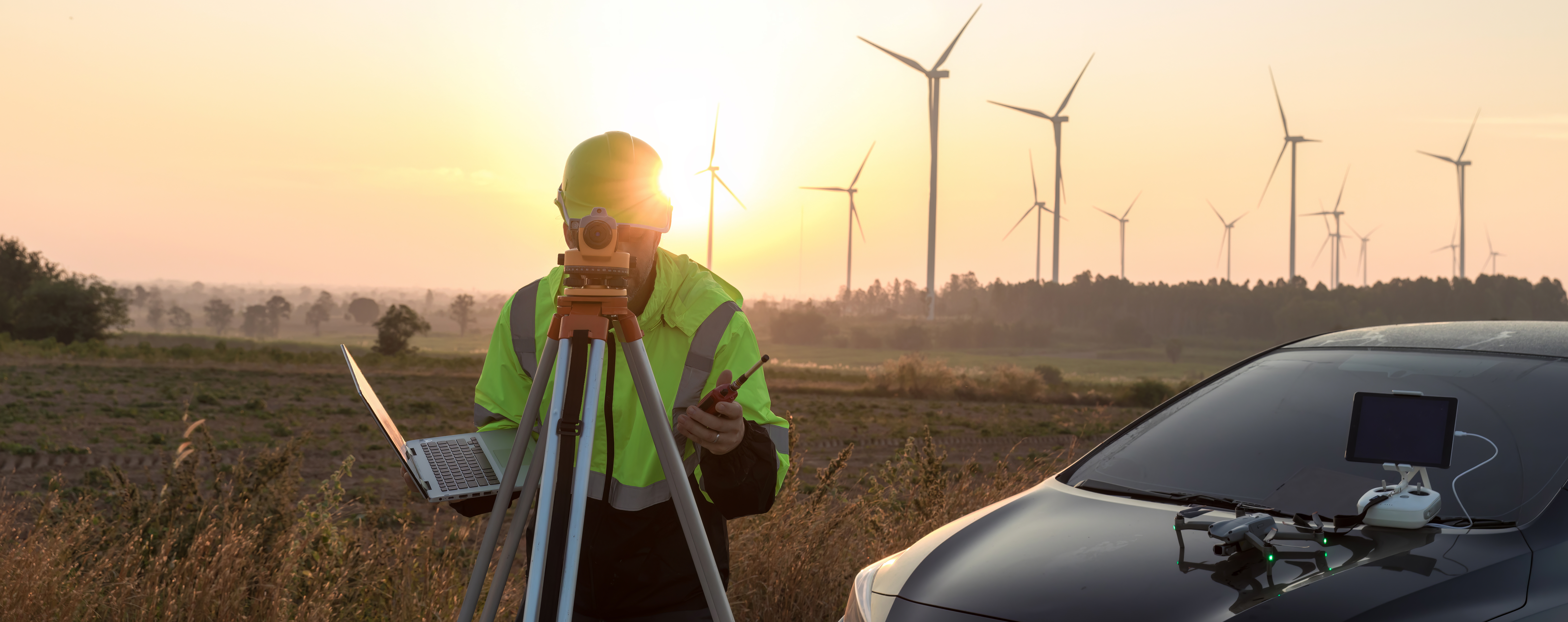 Zukunftsberufe - grün und digital Ein Vermessungsingenieur (mit Handy, Drohne und Laptop ausgestattet) untersucht die im Hintergrund befindlichen Windturbinen im Sonnenuntergang. Rechts im Bild befindet sich ein Auto.