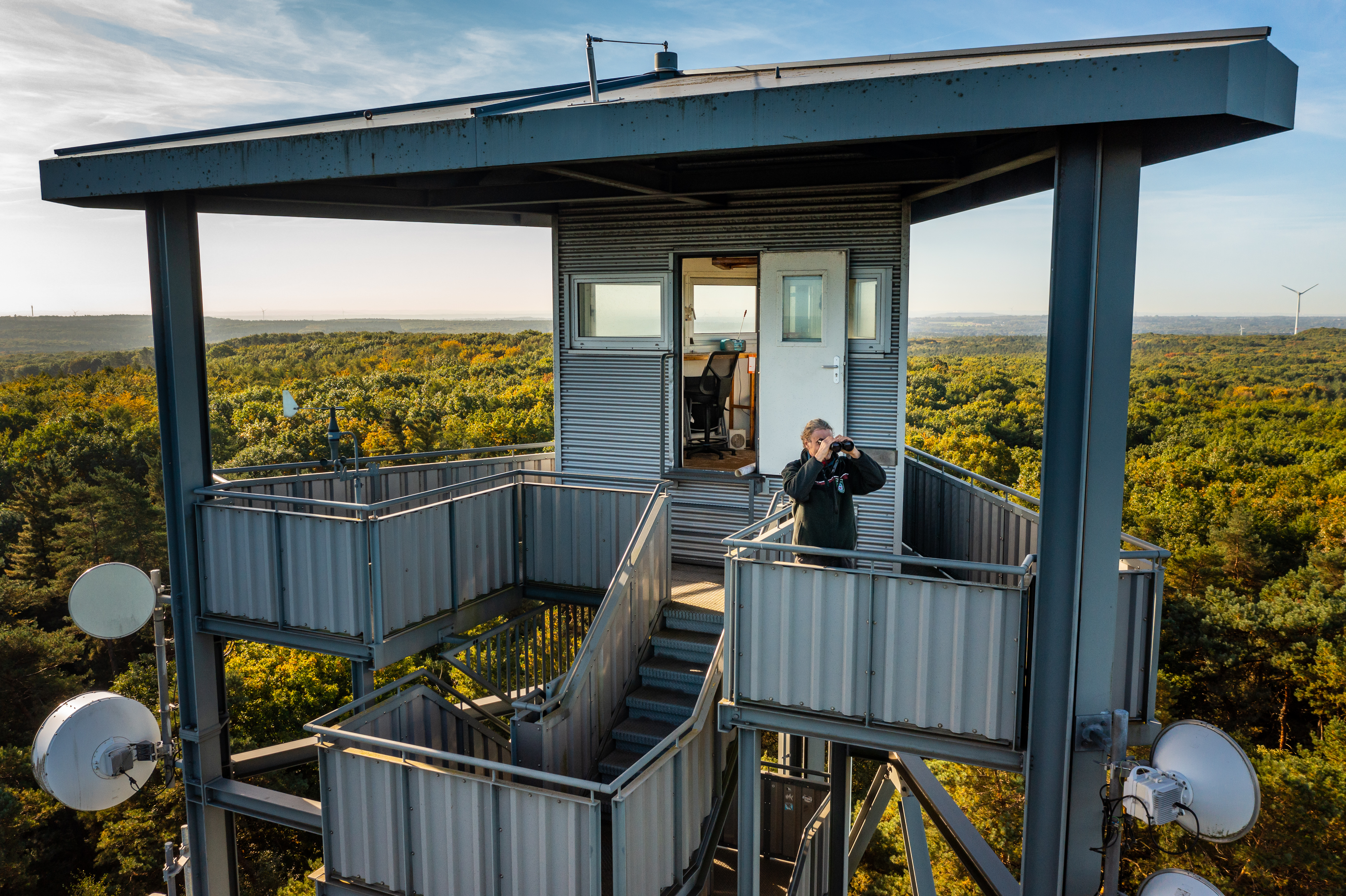 Revierleiter steht mit einem Fernglas auf einem Feuerwachturm umgeben von Wald.