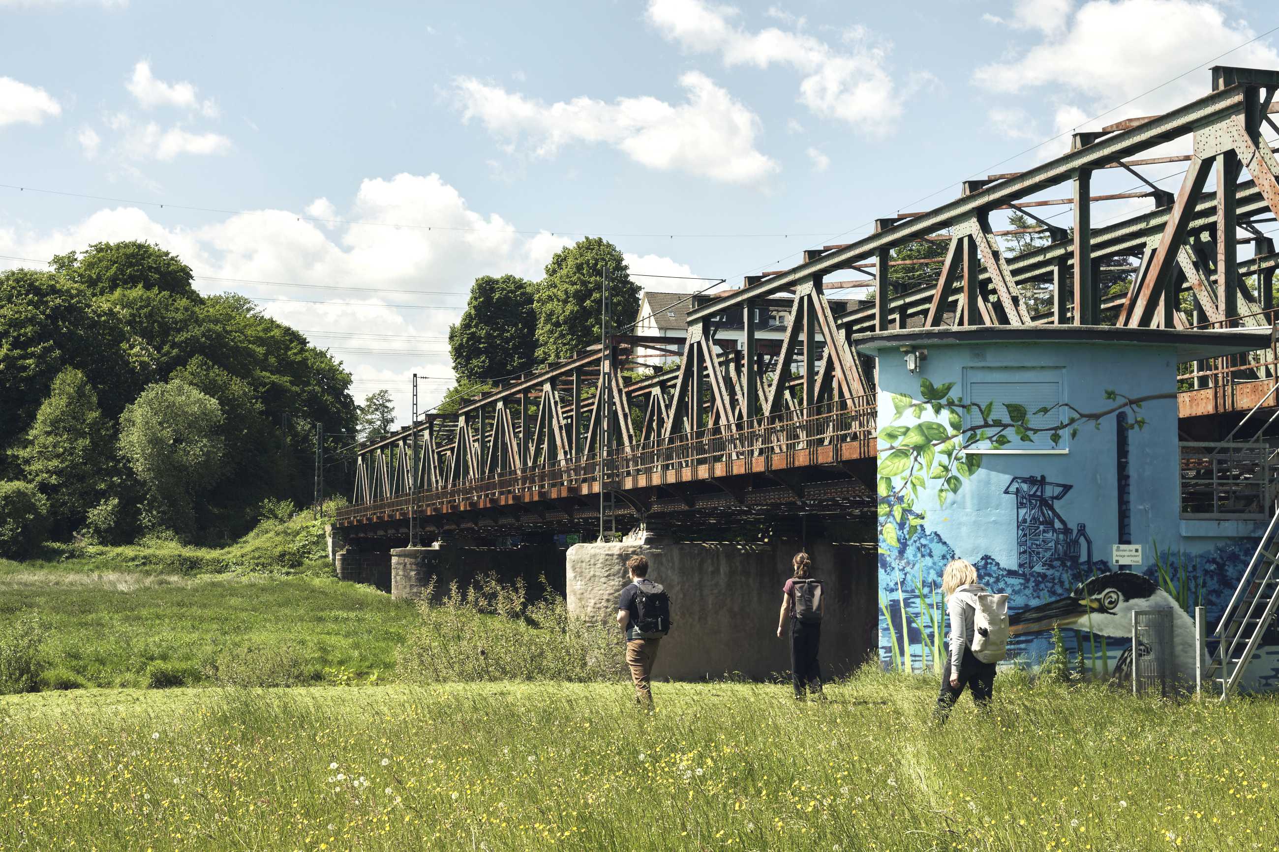 Drei Personen wandern auf einer grünen Wiese entlang einer Bahnstrecke.