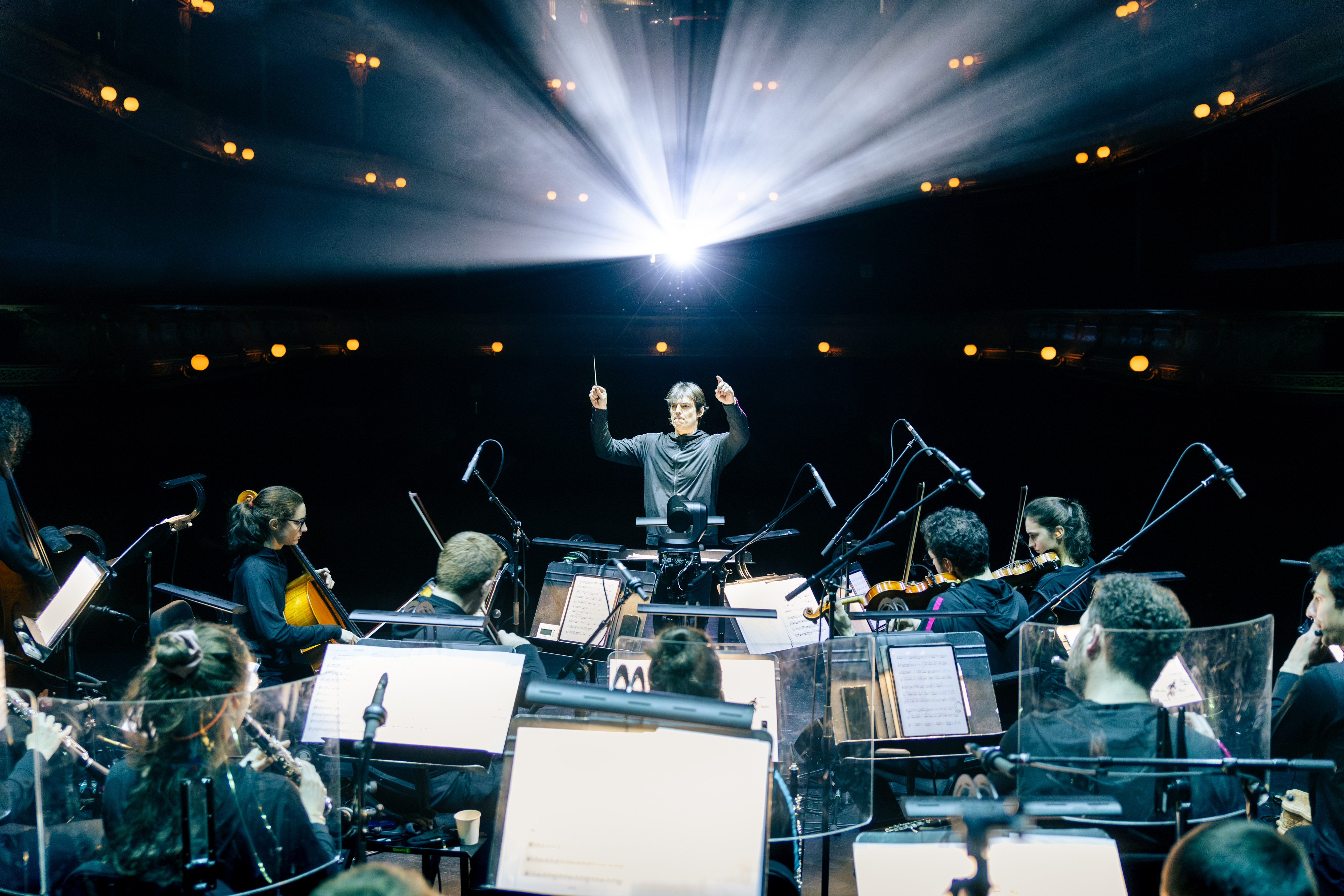 Julien Masmondet leitet das Ensemble Les Apaches ! auf der Bühne eines Theaters, während ein heller Lichtstrahl von hinten in den Saal strahlt.